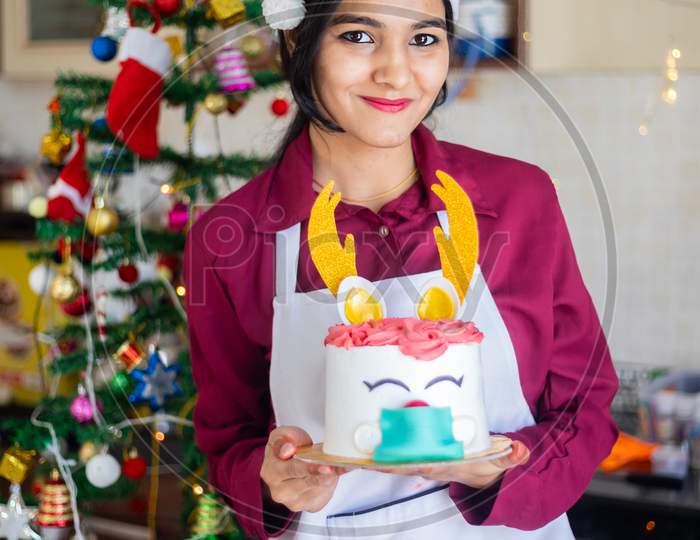 Portrait Young Smiling Indian Girl Wearing Apron And Santa Hat Holding Showing Cake At Home, Christmas Celebration During Covid-19 Pandemic Concept. Holidays And Festive Season.