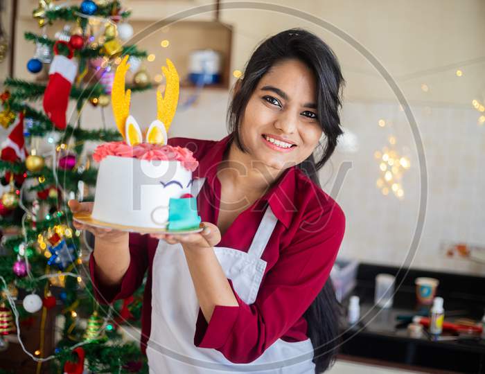 Portrait Young Happy Cheerful Indian Girl Wearing Apron Holding Showing Cake At Home, Christmas Celebration During Covid-19 Pandemic Concept. Holidays And Festive Season. Copy Space