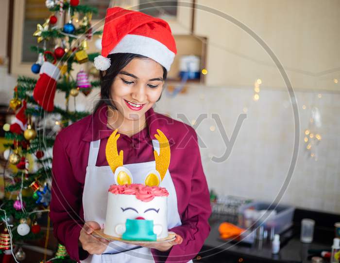 Portrait Young Smiling Indian Girl Wearing Apron And Santa Hat Holding Showing Cake At Home, Christmas Celebration During Covid-19 Pandemic Concept. Holidays And Festive Season.