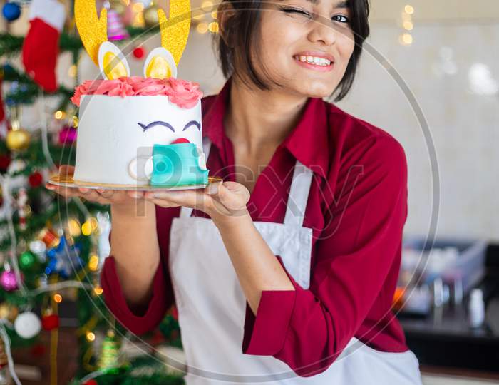 Portrait Young Cheerful Indian Girl Wearing Apron Holding Showing Cake At Home, Fun Christmas Celebration During Covid-19 Pandemic Concept. Holidays And Festive Season. Copy Space