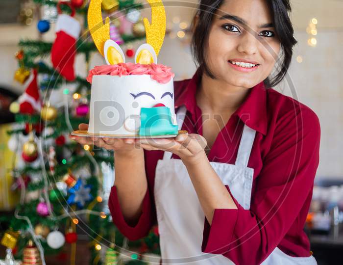 Portrait Young Happy Cheerful Indian Girl Wearing Apron Holding Showing Cake At Home, Christmas Celebration During Covid-19 Pandemic Concept. Holidays and Festive Season.