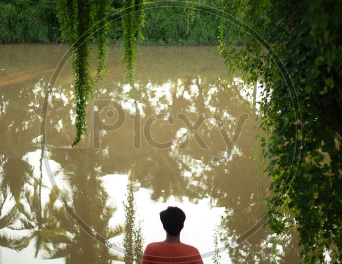 A Man  Standing At a Water Channel With Trees Reflection Over Water Surface at Rural Village Outskirts