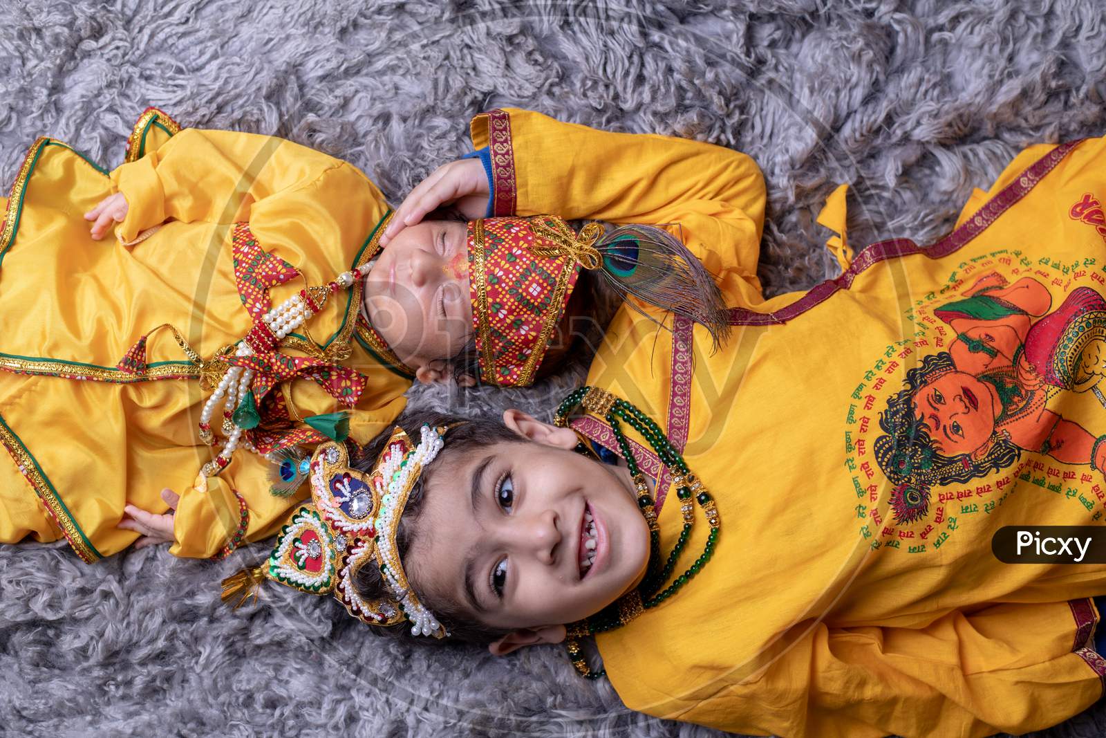 Young Indian Brothers Or Siblings Dressed Like Lord Sri Krishna And Posing Over A Gray Background