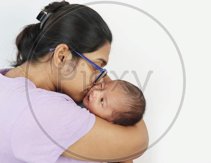 A Young Mother In Spectacles Kissing Her Infant Baby Boy In Her Lap In Solid Grey Background With Space For Text. Mothers Day Concept Photo.