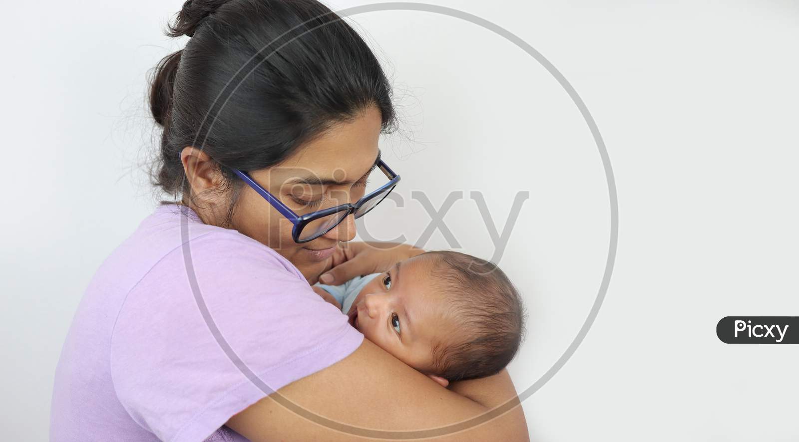 A Young Mother In Spectacles Looking At Her Infant Baby Boy In Her Lap In Solid Grey Background With Space For Text. Mothers Day Concept Photo.