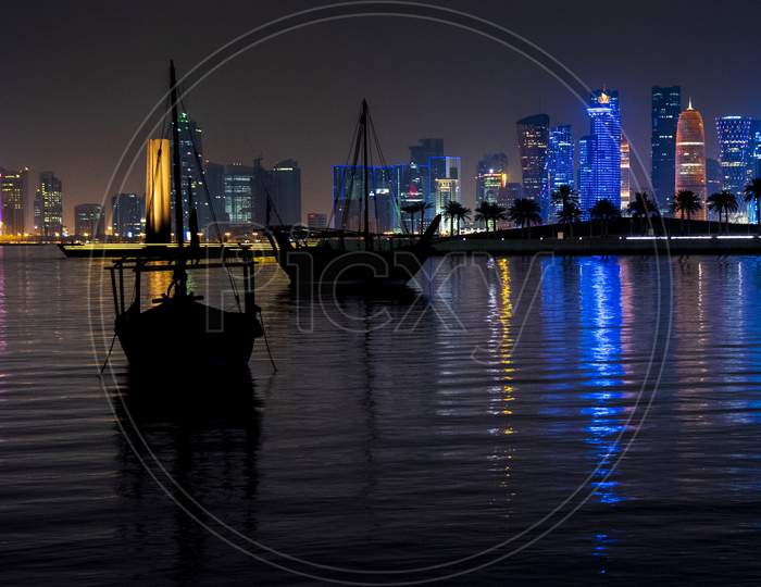 Silhouette Of Boats On Sea At West Bay In Doha With Urban City Buildings In Night Lights effect