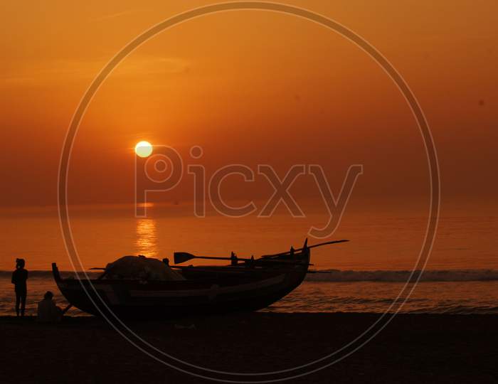 Silhouette Of Lone Fishing Boat Over Sea And Sunset Sky In Background