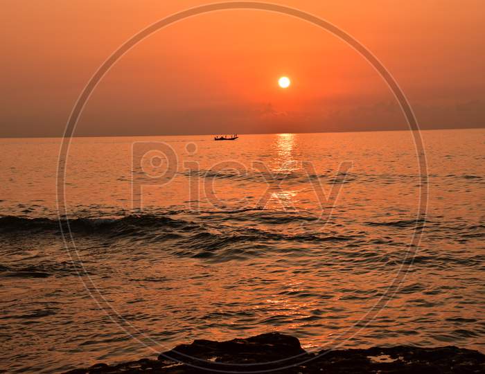 Silhouette Of Lone Fishing Boat Over Sea And Sunset Sky In Background