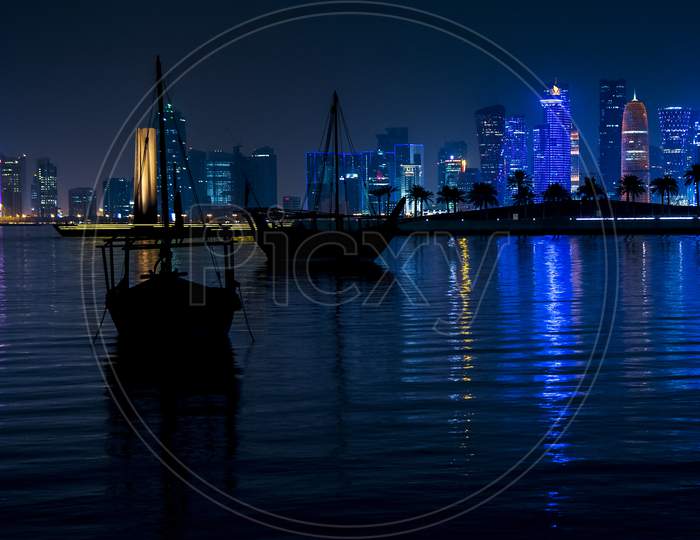 Silhouette Of Boats On Sea At West Bay In Doha With Urban City Buildings In Night Lights effect