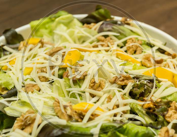 Closeup Of Delicious Salad Of Lettuce, Watercress, Arugula, Mango, Walnuts And Shredded Cheese On White Ceramic Platter On Wooden Table. Healthy And Nutritious Food. Selective Focus.