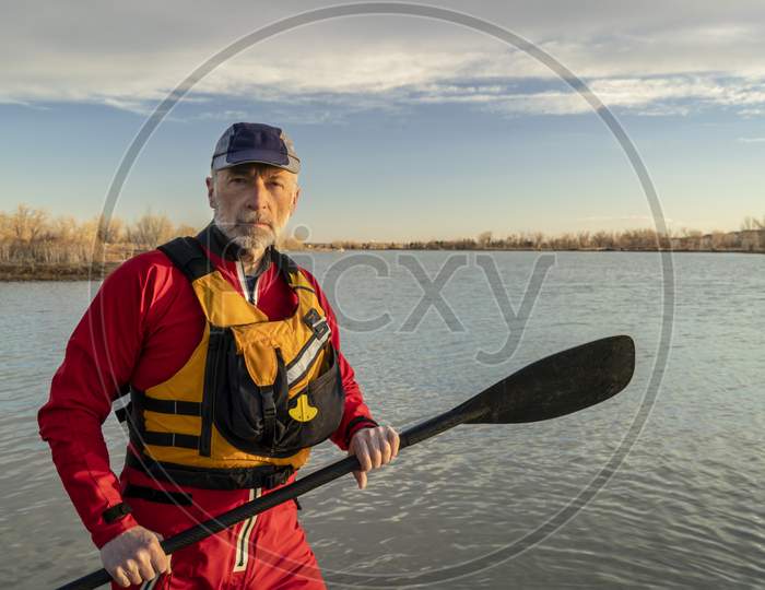 Environmental Portrait Of A Senior Male Paddler In A Drysuit And Life Jacket Holding Stand Up Paddle On A Shore Of A Lake