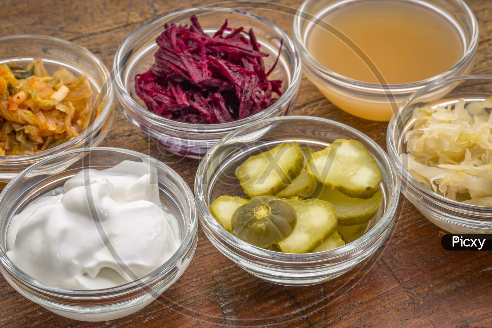 A Sampler Of Fermented, Prebiotics, Food Great For Gut Health - Glass Bowls Against Wood:  Kimchi, Red Beets, Apple Cider Vinegar, Coconut Milk Yogurt, Cucumber Pickles, Sauerkraut