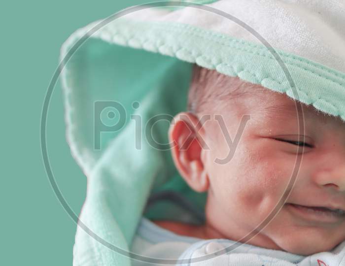 A Smiling Newborn Baby With Dimple In Cheek Wrapped In Sea Green Colored Towel With Hood With Eyes Closed With Selective Focus On Front Eye