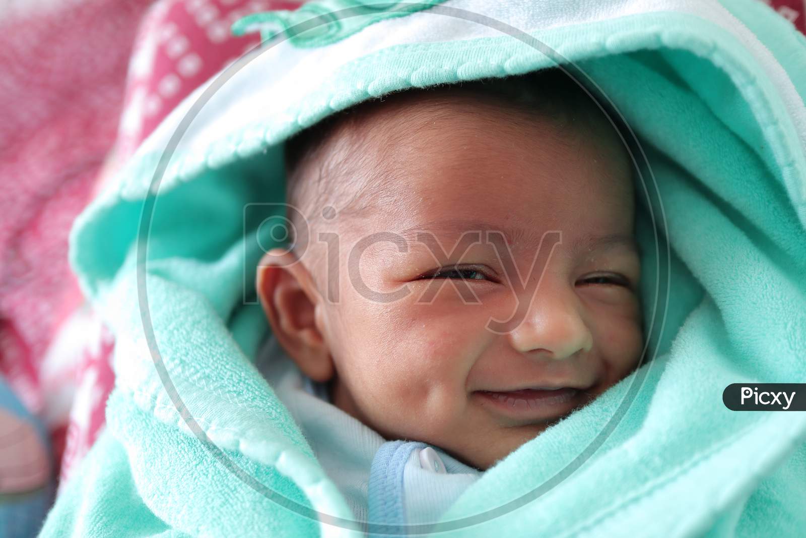 A Smiling Newborn Baby With Dimple In Cheek Wrapped In Sea Green Colored Towel With Hood With Eyes Closed With Selective Focus On Front Eye.