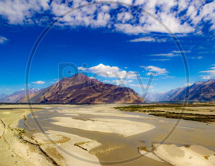 High Dynamic Range Image Of Barren Mountain In A Desert With River And Deep Blue Sky And White Patchy Clouds In Ladakh, Jammu And Kashmir, India