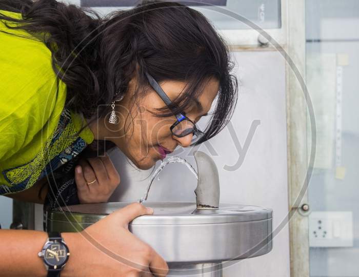 An Indian Lady In Green Dress Drinking Water From A Steel Water Fountain On A Sink By Pressing Button At Airport