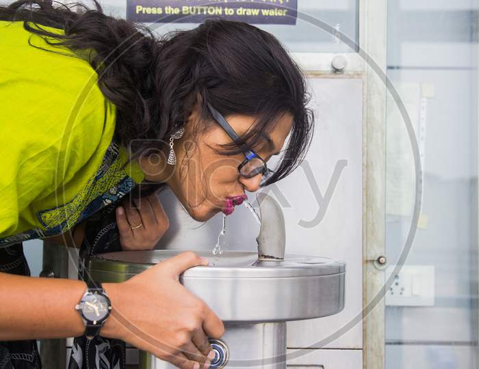 An Indian Lady In Green Dress Drinking Water From A Steel Water Fountain On A Sink By Pressing Button At Airport