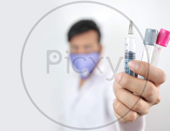 A Medical Professional In White Coat And Protective Mask Holding Vials And Syringe In Hand With Selective Focus In Hand With Blurred Background.