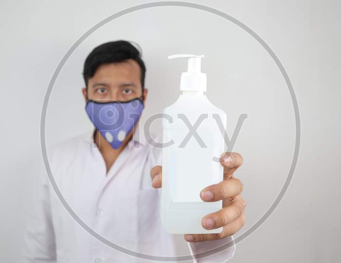 A Male Doctor In Mask And White Coat Holding A White Mockup Bottle Of Hand Sanitizer Isolated In Grey Background With Space For Text.