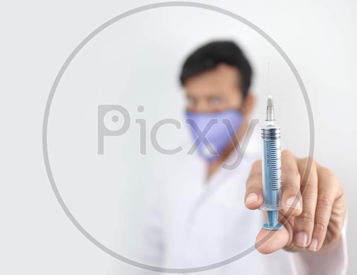A Medical Professional In White Coat And Protective Mask Holding Syringe In Hand With Selective Focus In Hand With Blurred Background.