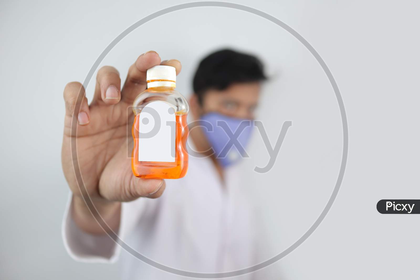 A Male Doctor In Mask And White Coat Holding A White Mockup Bottle Of Anti Septic Isolated In Grey Background With Space For Text.