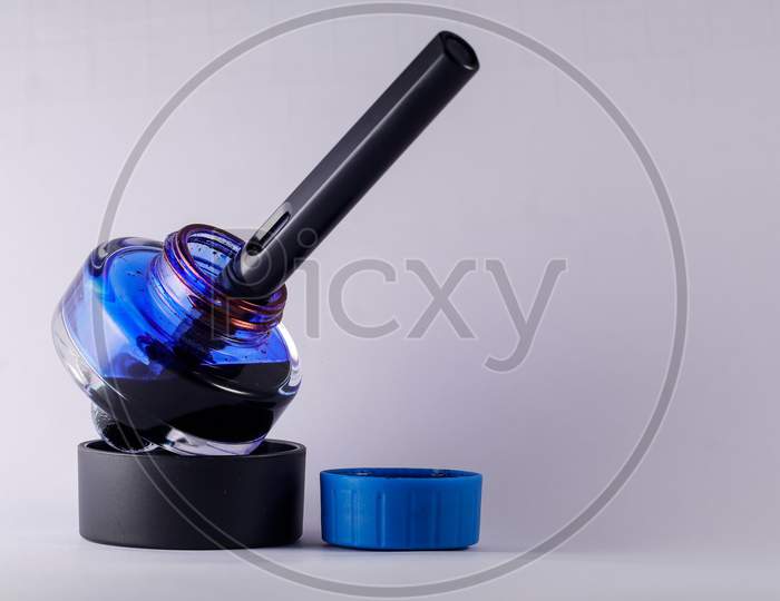 A View Of Open Blue Ink Bottle With A Fountain Pen Dip Inside Against White Background