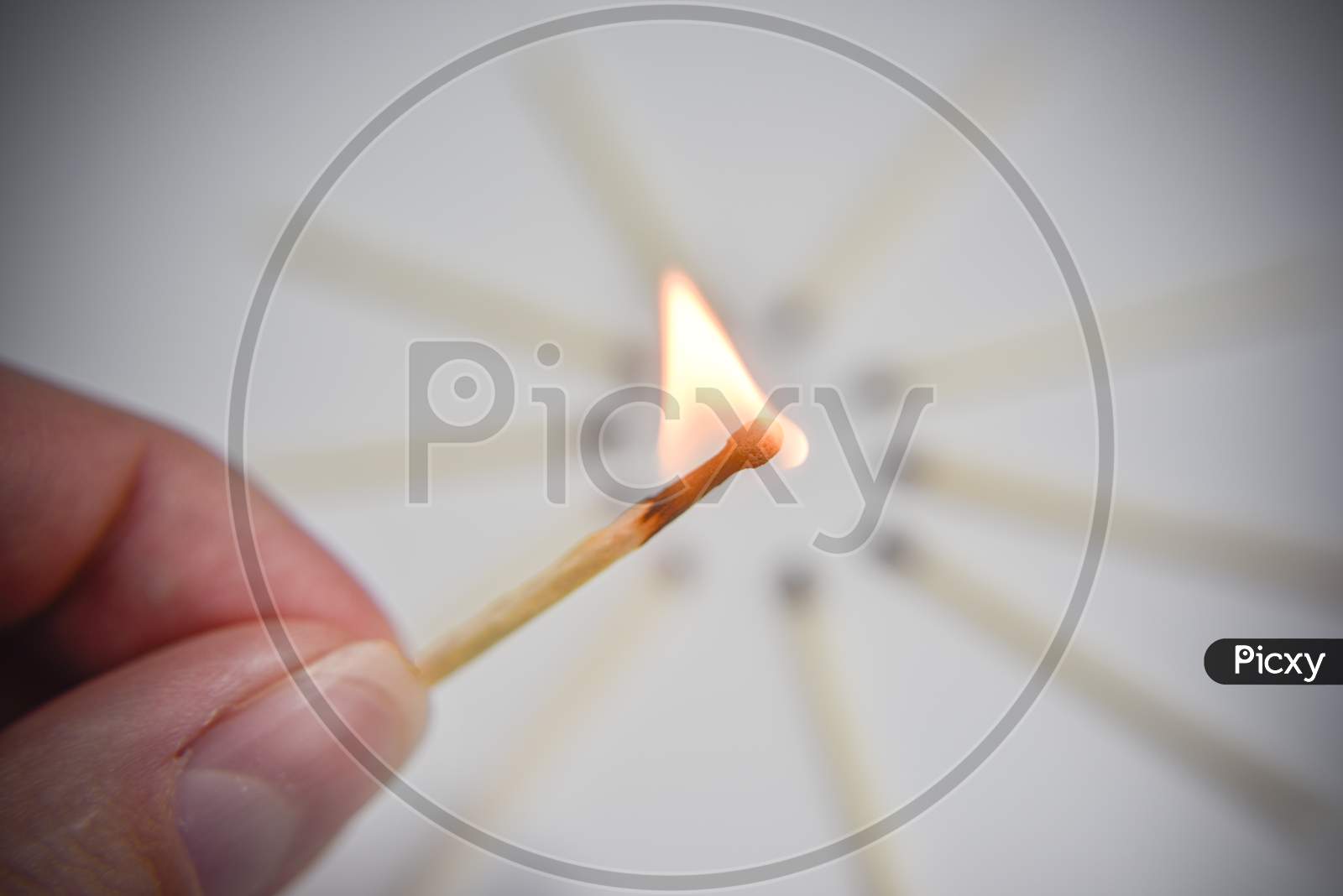 Burning Wooden Match In The Hand With Circle Of Matches On The White Backgtound. Selective Focus. Chain Reaction Concept.