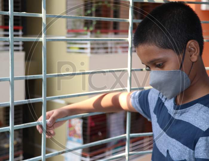 Passing Boring Time Due To Corona Virus Outbreak. Lock Down And School Closures. A School Boy With Face Mask Watching Outside Through The Window. Covid-19 Pandemic Now In The World.