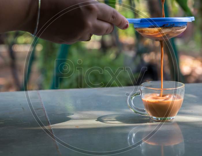 A Hand Pouring Steamy Masala Tea Through Plastic Strainer Into A Glass Cup