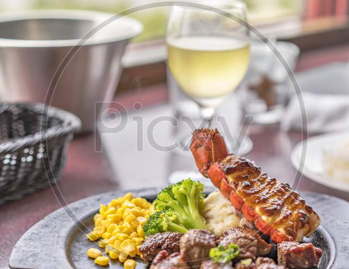 Beef Steack And Seafood Lobster Tail With Cheese, Corn And Broccoli On Plate With White Wine In Glass.