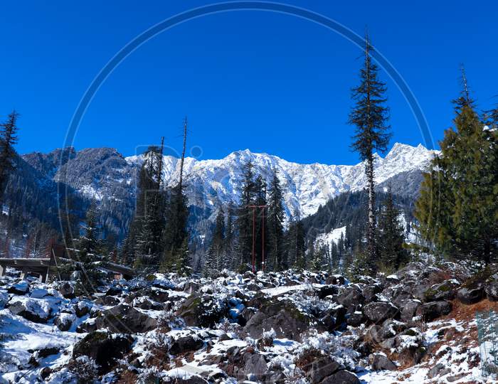 Snow-Covered Mountains With Pine Trees In The Background And Rocks Covered With Snow In Front.