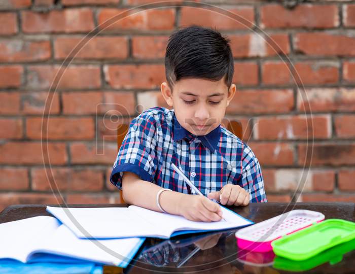 Cute Little Male Student In School Uniform Writing On Notebook While Attending Class In Primary School. He Is Using Pencil For Writing.