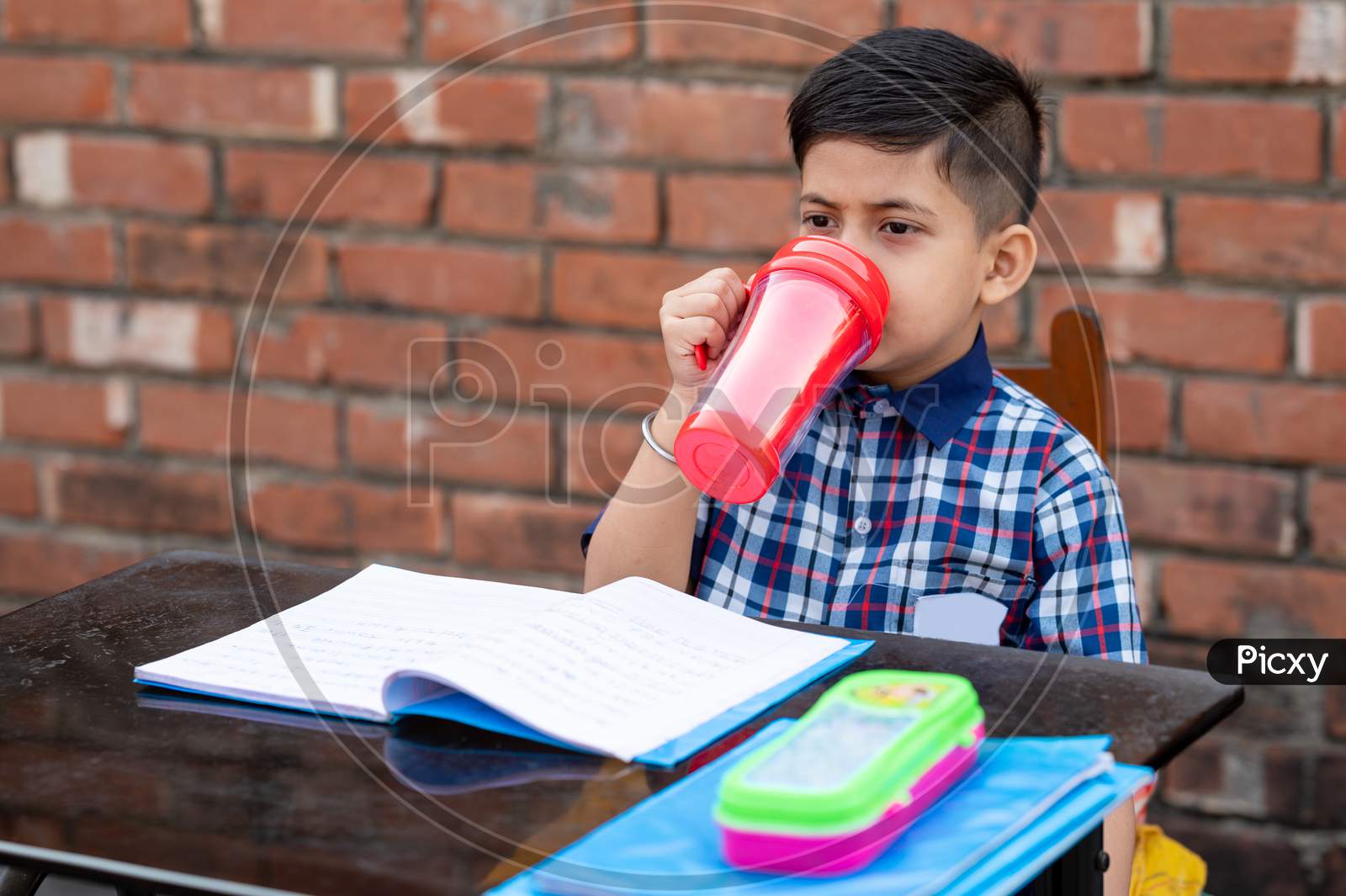 Primary School Student Drinking Water With Red Water Bottle While Sitting In Class On Study Table. Indian School Student In Classroom.