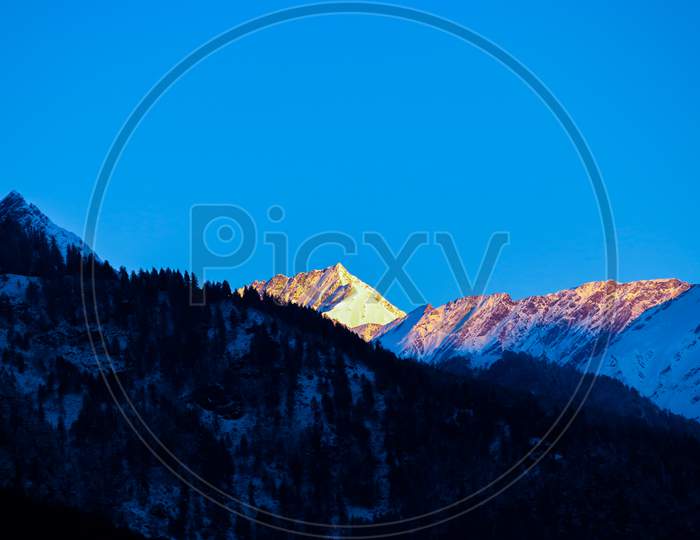 Sunrise On The Top Of A Snow-Covered Mountain. The Mountain In The Front Is Covered With Pine Trees.