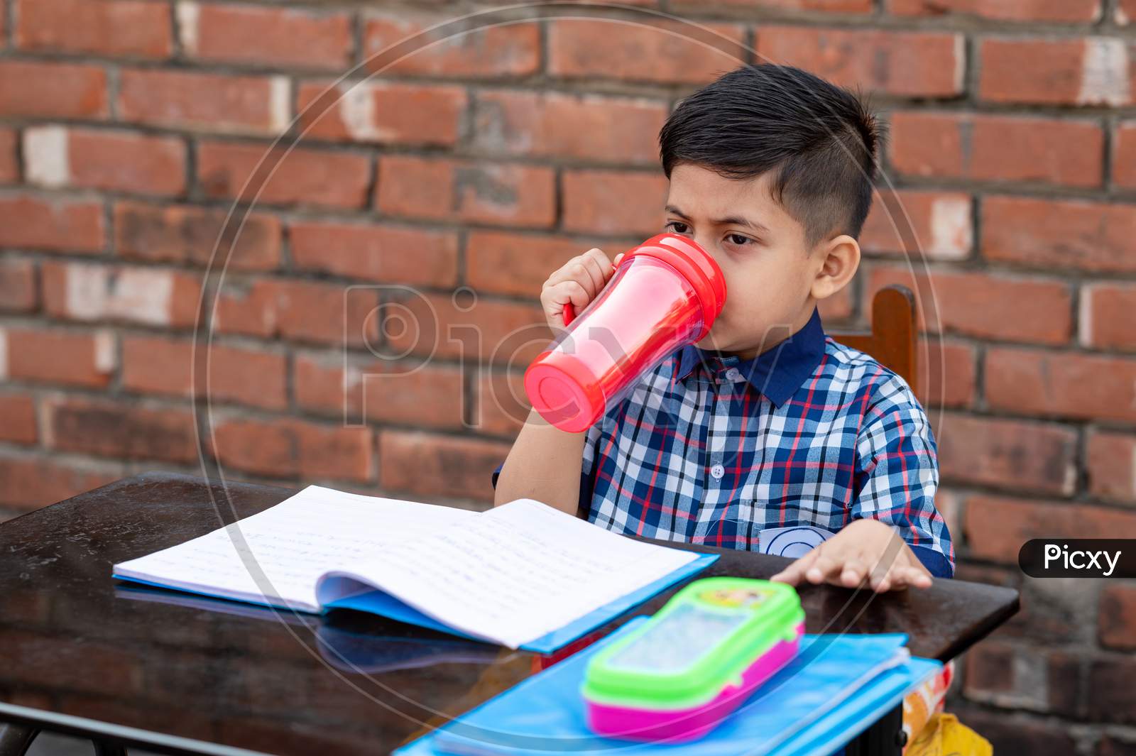 Primary school student drinking water with red water bottle while sitting in class on study table. Indian school student in classroom.