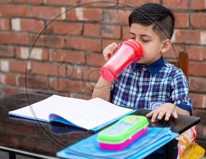 Primary School Student Drinking Water With Red Water Bottle While Sitting In Class On Study Table. Indian School Student In Classroom.