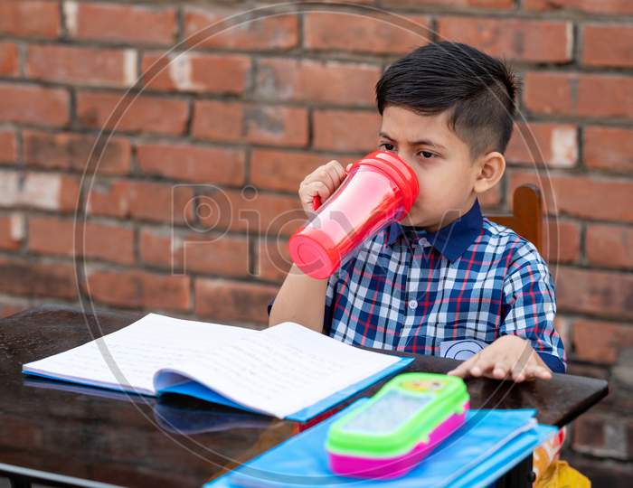 Primary school student drinking water with red water bottle while sitting in class on study table. Indian school student in classroom.