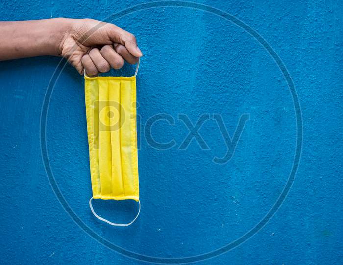 Man Holding Yellow Face Mask Holding On His Hand Isolated On Blue Background. Protective Face Mask On Hand Against Covid-19. Color Face Mask.Space For Text