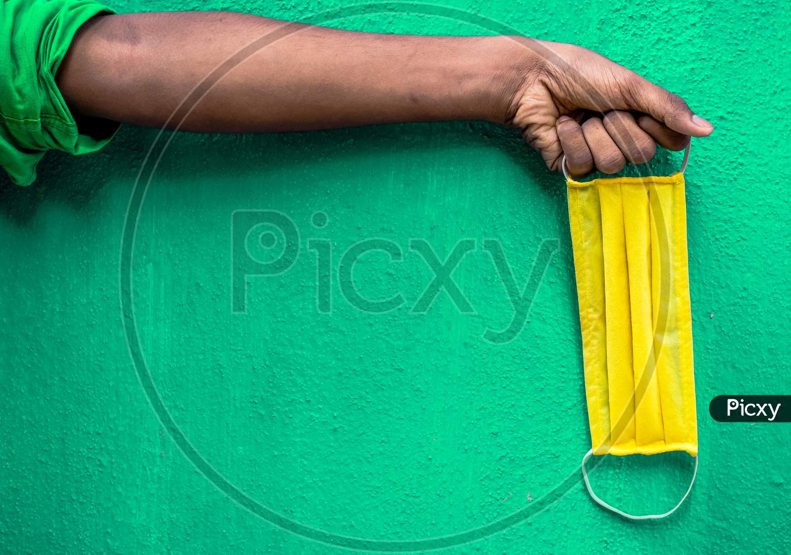 Man Holding Yellow Face Mask Holding On His Hand Isolated On Green Background. Protective Face Mask On Hand Against Covid-19. Color Face Mask.Space For Text