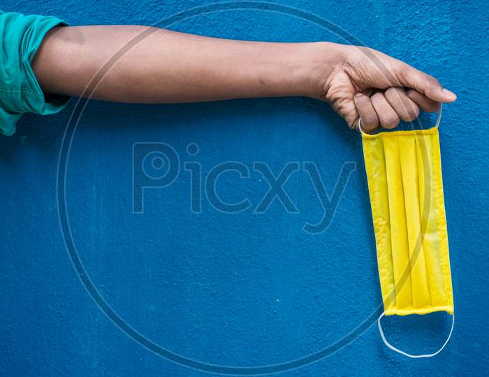 Man Holding Yellow Face Mask Holding On His Hand Isolated On Blue Background. Protective Face Mask On Hand Against Covid-19. Color Face Mask. Space For Text