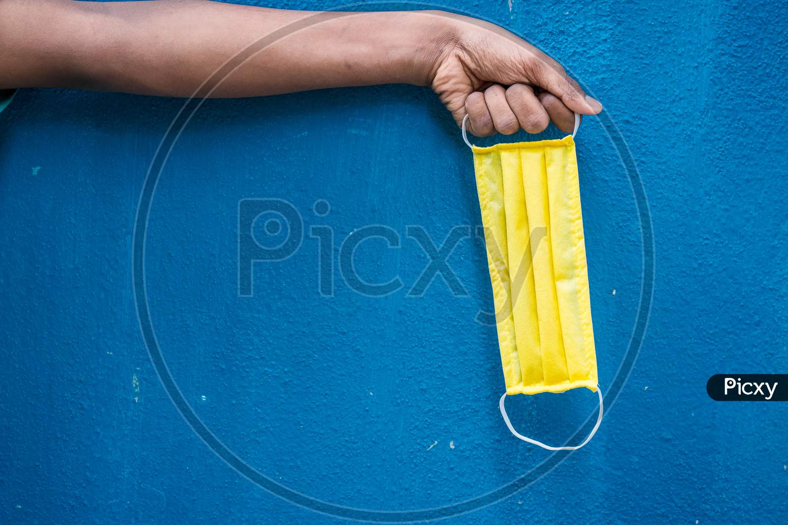 Man Holding Yellow Face Mask Holding On His Hand Isolated On Blue Background. Protective Face Mask On Hand Against Covid-19. Color Face Mask.Space For Text