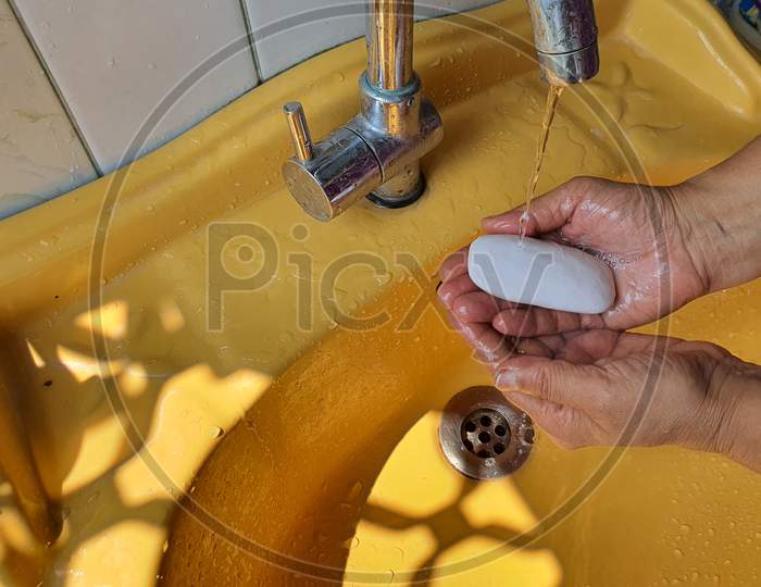 Washing Of Both Hands With Soap Under A Yellow Wash Basin With Running Tap Water.