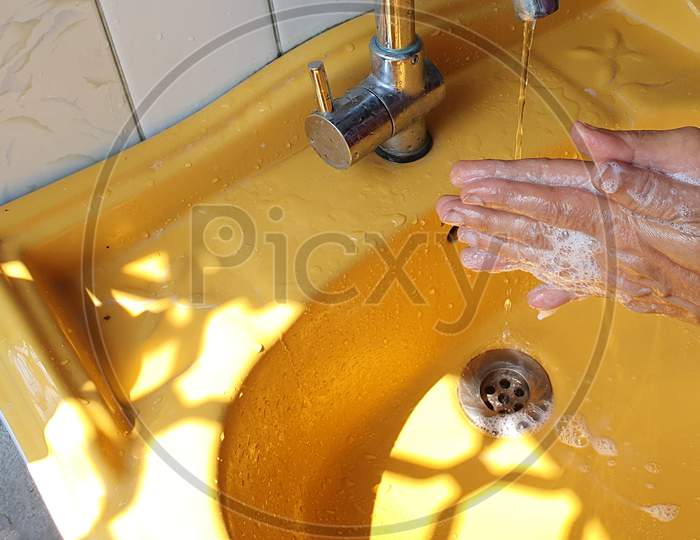 Washing Of Both Hands With Soap Under A Yellow Wash Basin With Running Tap Water To Produce Foam And Lather.