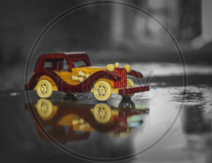 Reflection Vintage Toy Car Under The Rain On The Road. Miniature Car Toy Reflection In Rain. Reflection Red And Yellow Vintage Car On Rain. Rain Falling On Vintage Car Toy Close
