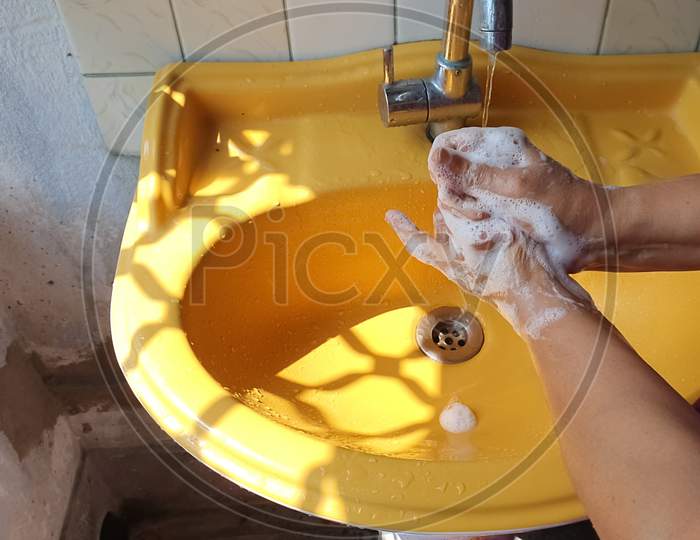 Washing Of Both Hands With Soap Under A Yellow Wash Basin With Running Tap Water To Produce Foam And Lather.
