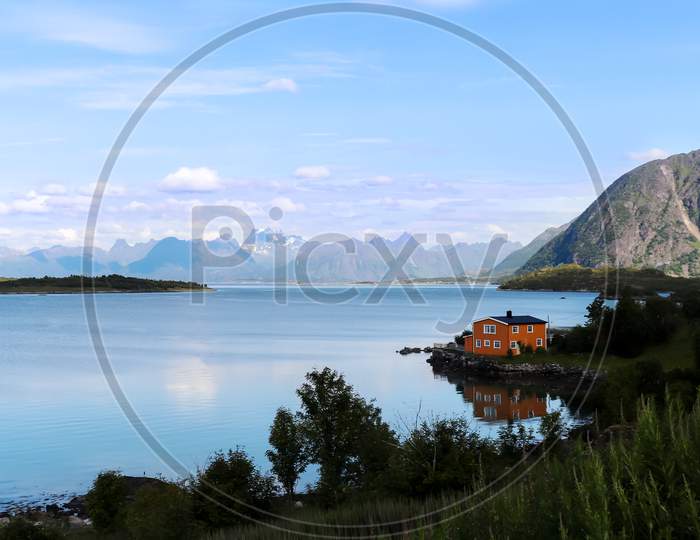 Orange House On The Edge Of A Blue Lake With Blue Mountains In The Background On The Lofoten Islands In Norway.