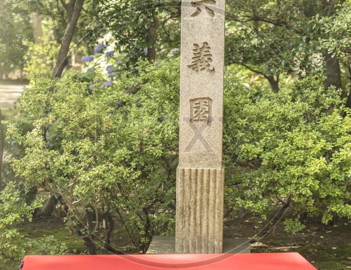 Rectangular Stone Testifying The Status Of Japanese National Scenic Site In The Rikugien Garden In Tokyo. Wooden Benches Covered With Red Felt Are Available To The Public To Enjoy A Moment Of Relaxation In The Park.