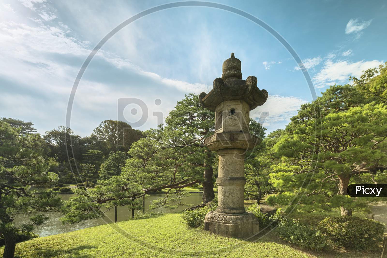 Giant Stone Lantern Around A Pond Surrounded By Pine Tree Under The Blue Sky In The Garden Of Rikugien In Tokyo In Japan.