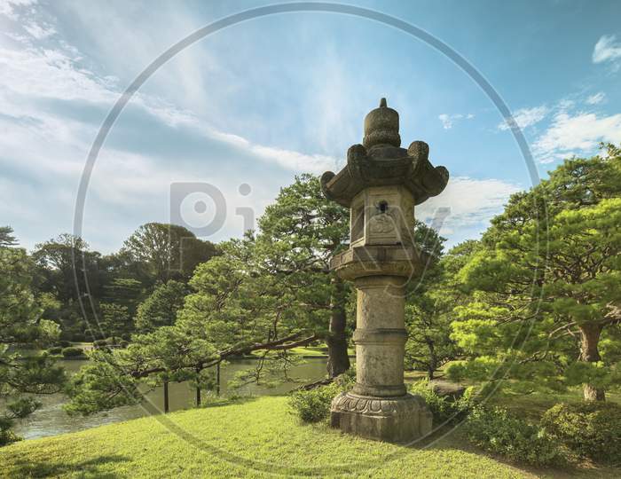 Giant Stone Lantern Around A Pond Surrounded By Pine Tree Under The Blue Sky In The Garden Of Rikugien In Tokyo In Japan.