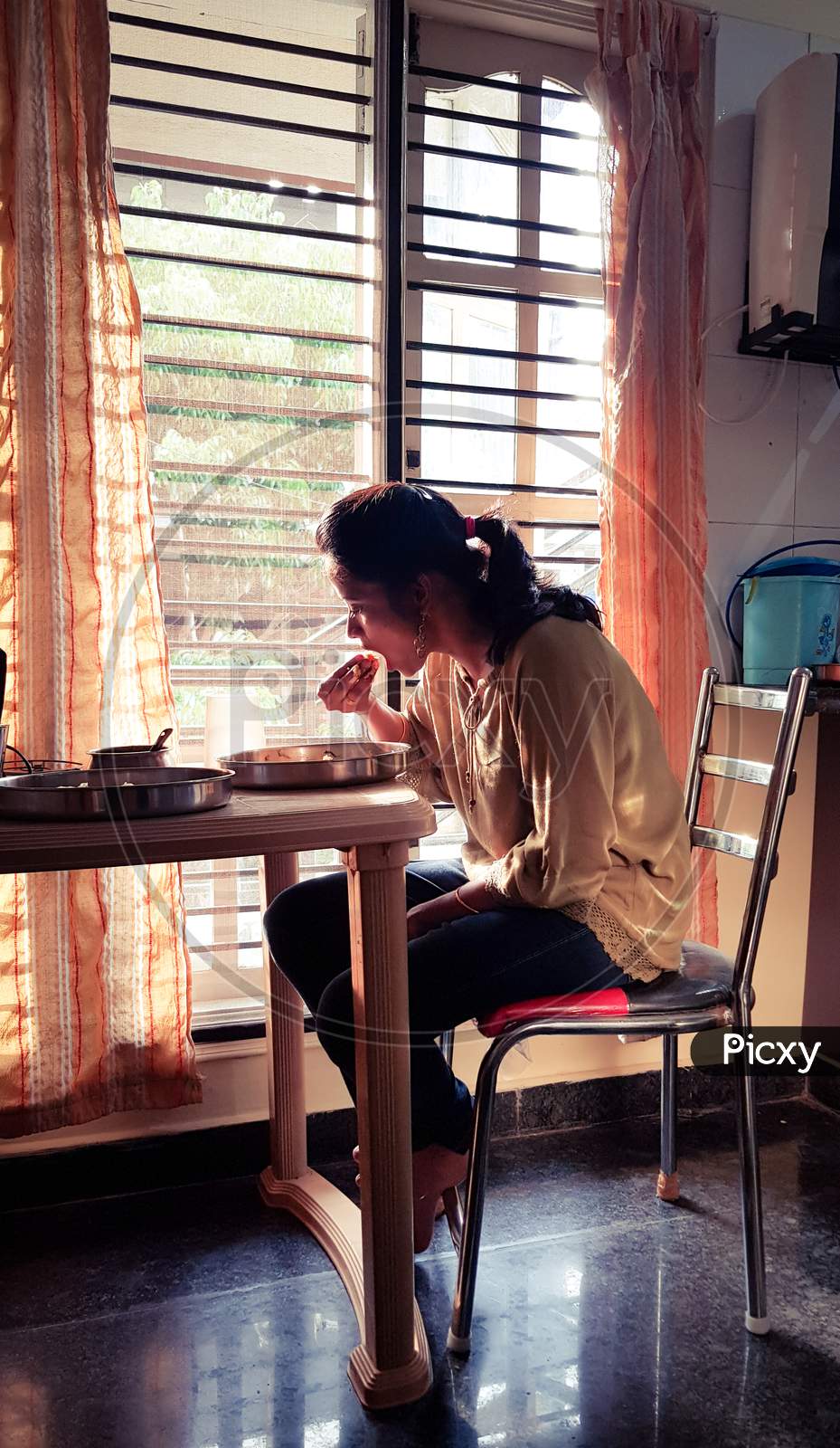 Bengaluru, Karnataka / India - April 26 2019: A young woman sitting by the window during morning hours and eating food from a steel plate